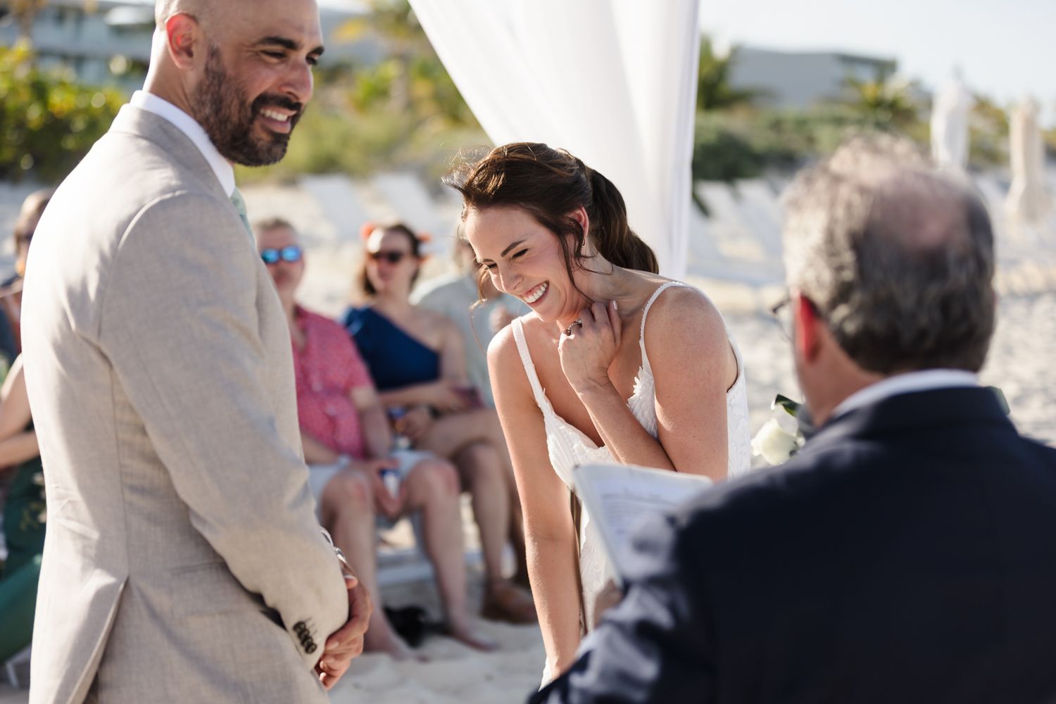 Bride and groom share a joyful moment during an outdoor beach wedding ceremony with guests seated in the background.