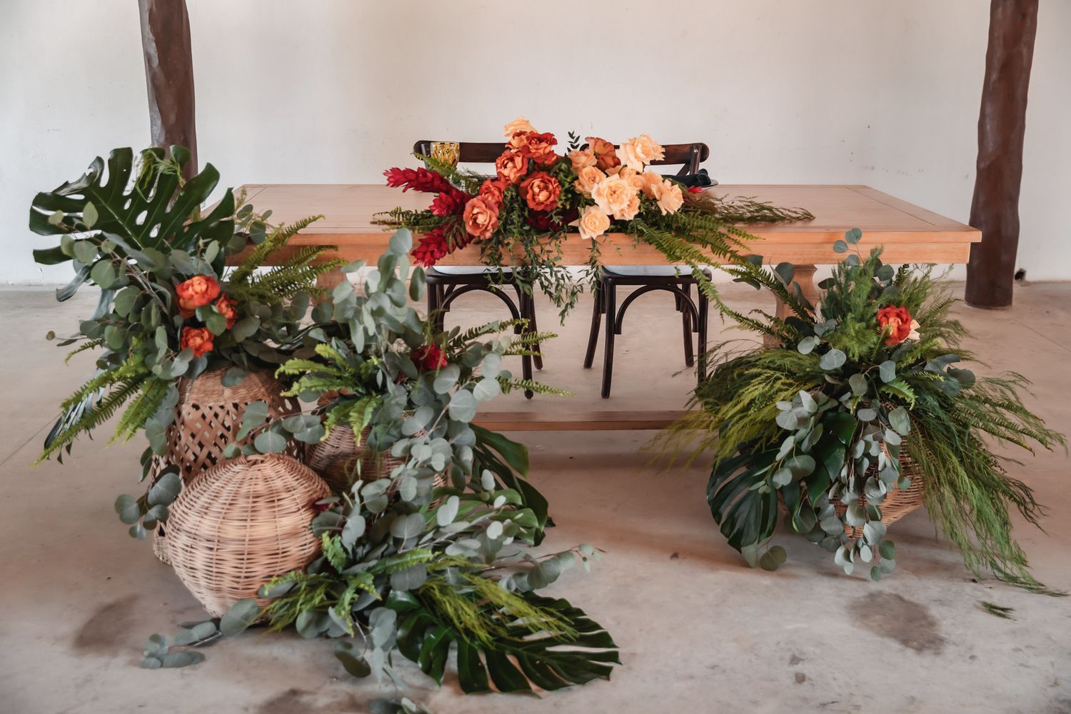 Rustic sweetheart table decorated with lush tropical greenery and orange floral arrangements on a barn floor.