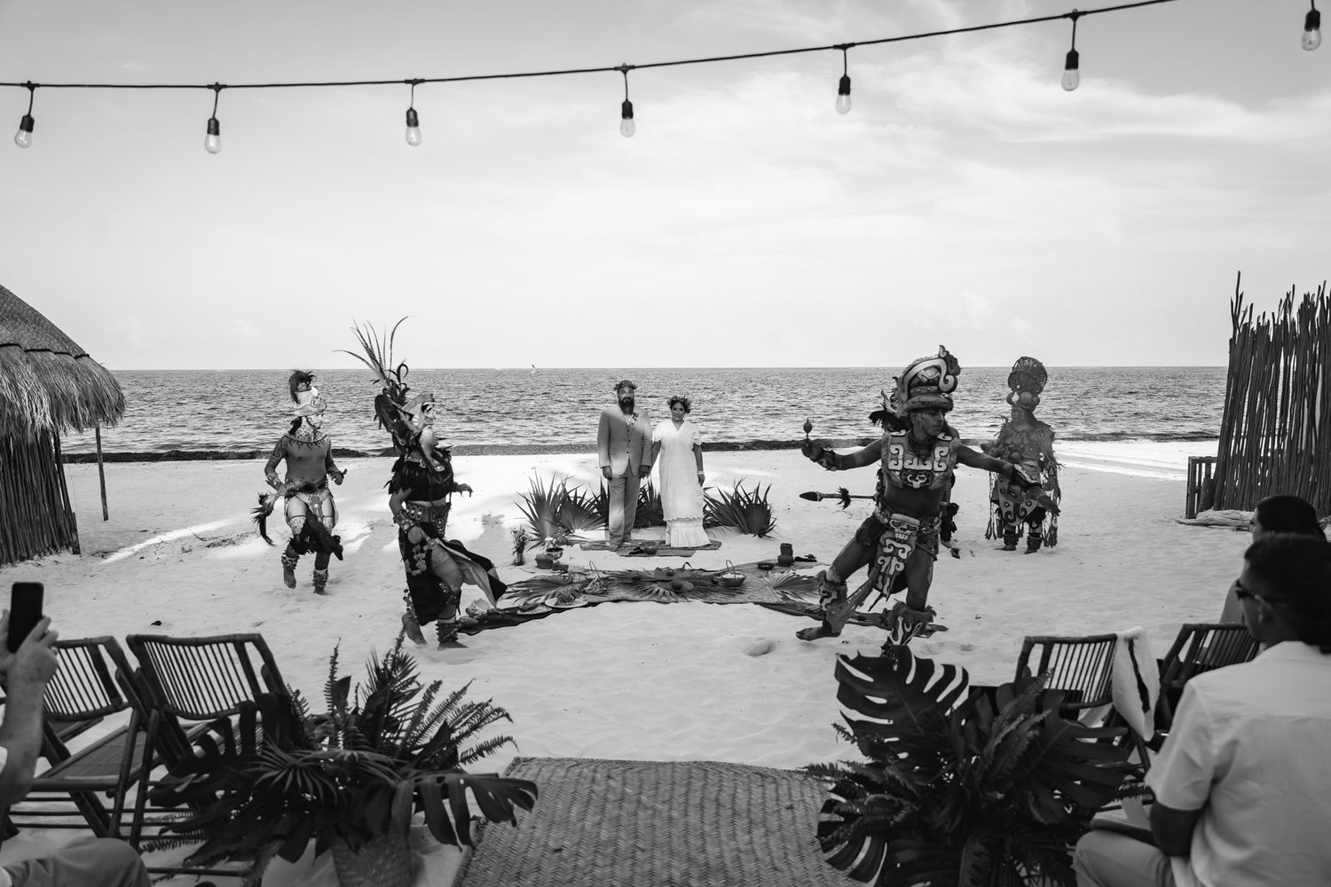 Traditional dancers perform on a beachside stage with ocean views in a black and white photo.