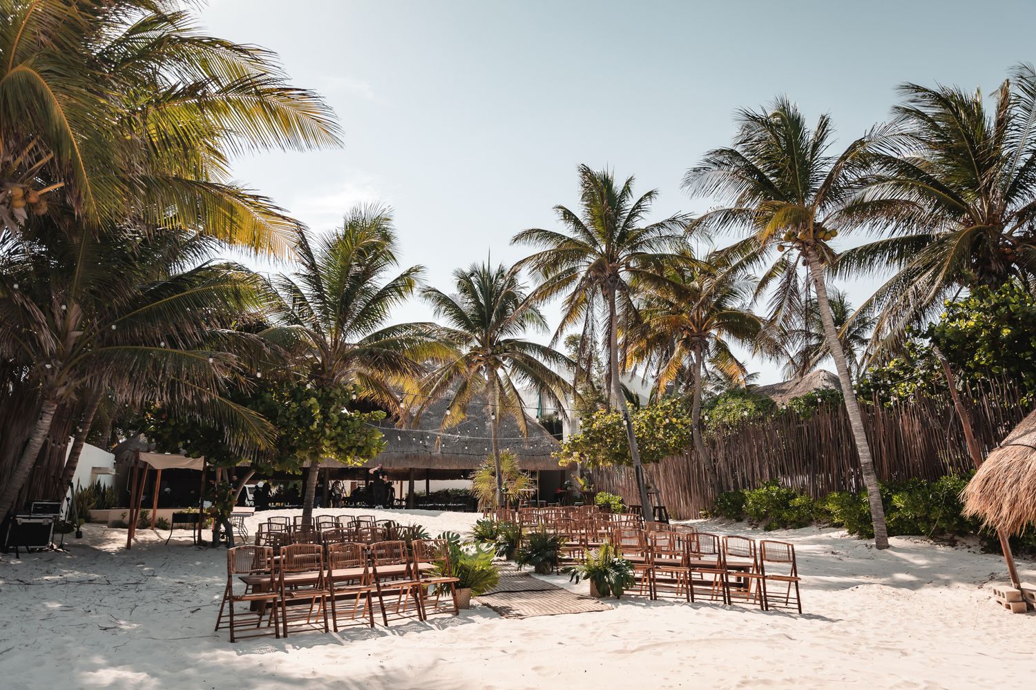 Outdoor beach wedding ceremony setup with wooden chairs on white sand surrounded by tall palm trees in a tropical setting.