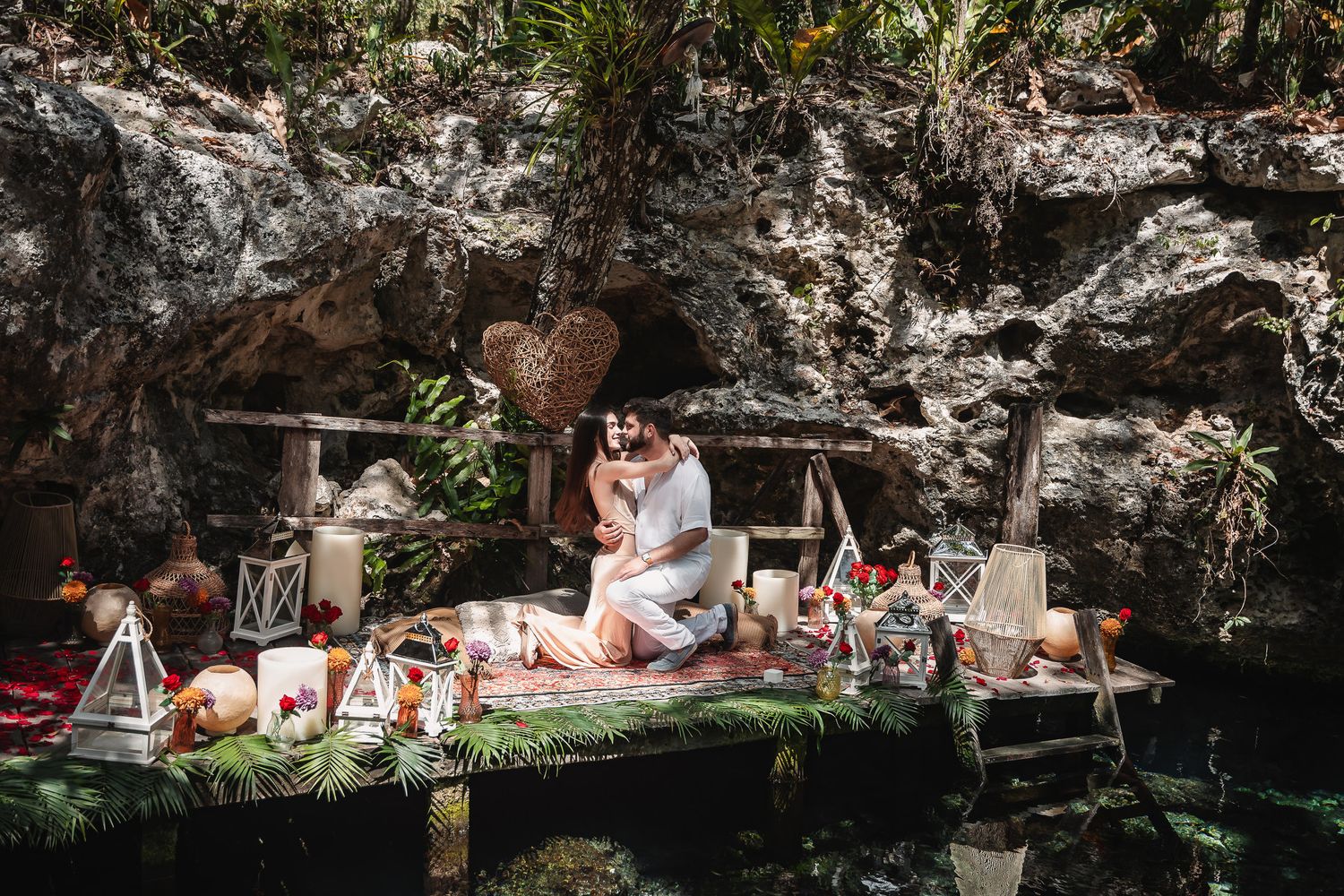 Couple sharing romantic moment on decorated jungle platform with candles, flowers, and tropical decor near rocky cave.
