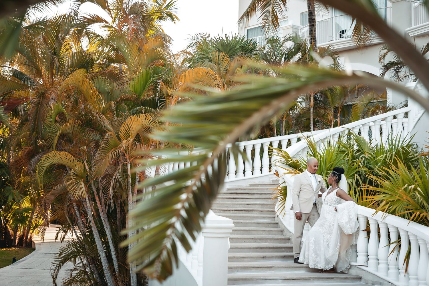 Bride and groom walk up white staircase surrounded by lush tropical palm trees at an elegant outdoor wedding venue.