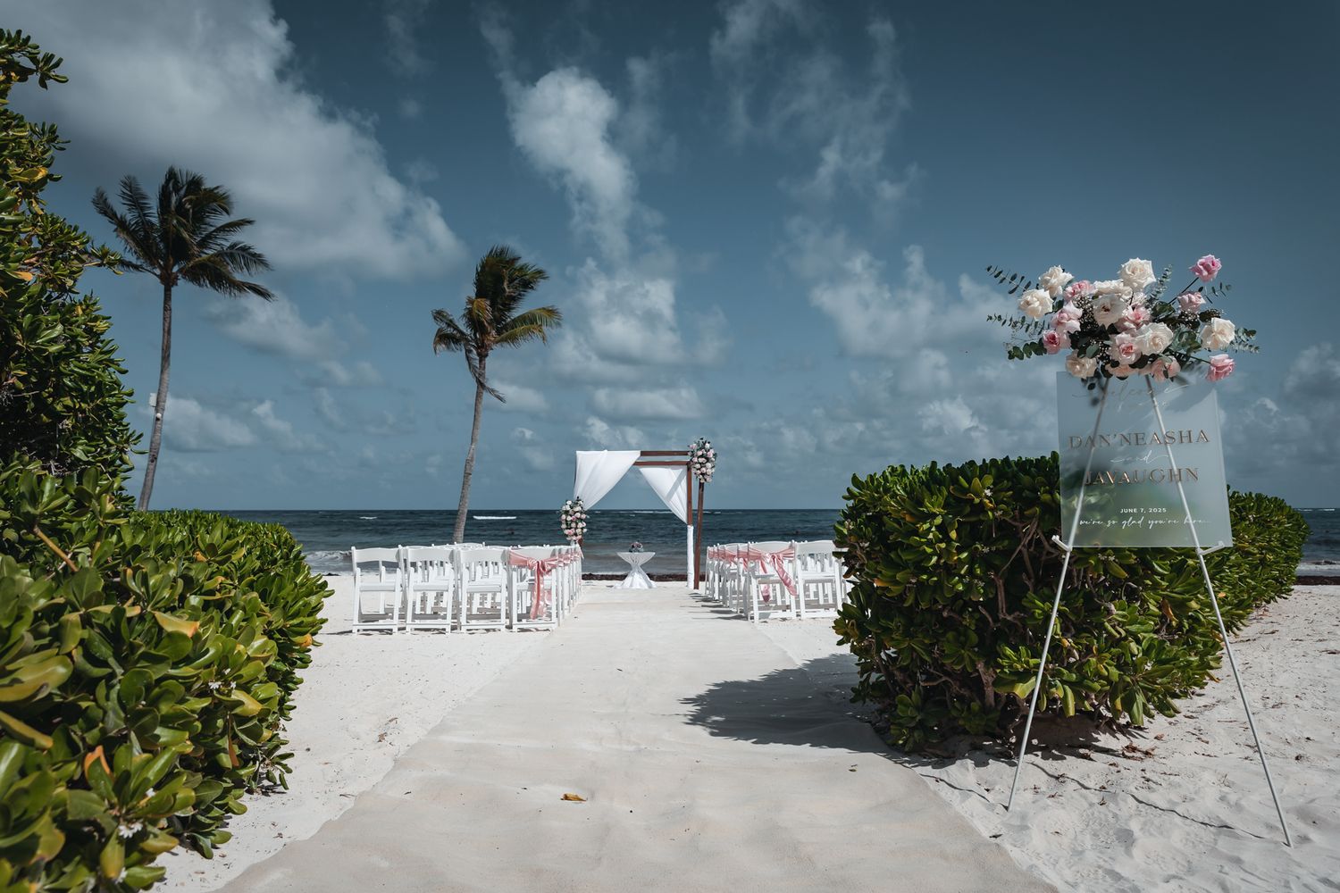 Beach wedding setup with white chairs, floral arch, and pink roses on sandy shore with palm trees and ocean backdrop.