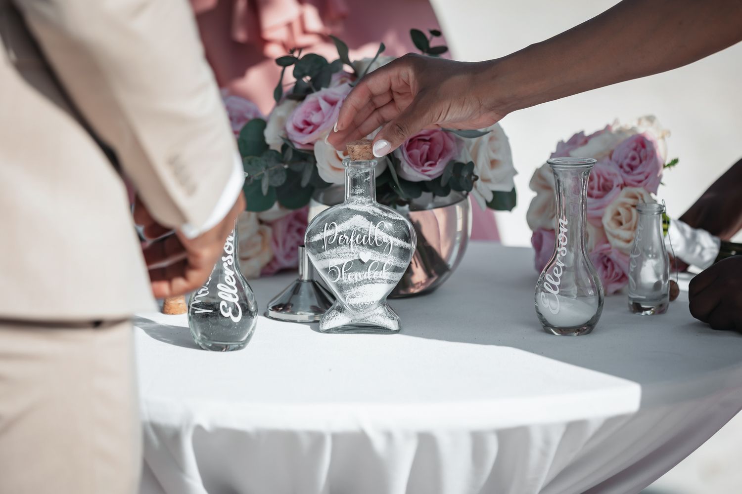 Hands pouring sand into a decorative wedding unity sand ceremony vase on a white table with pink roses.
