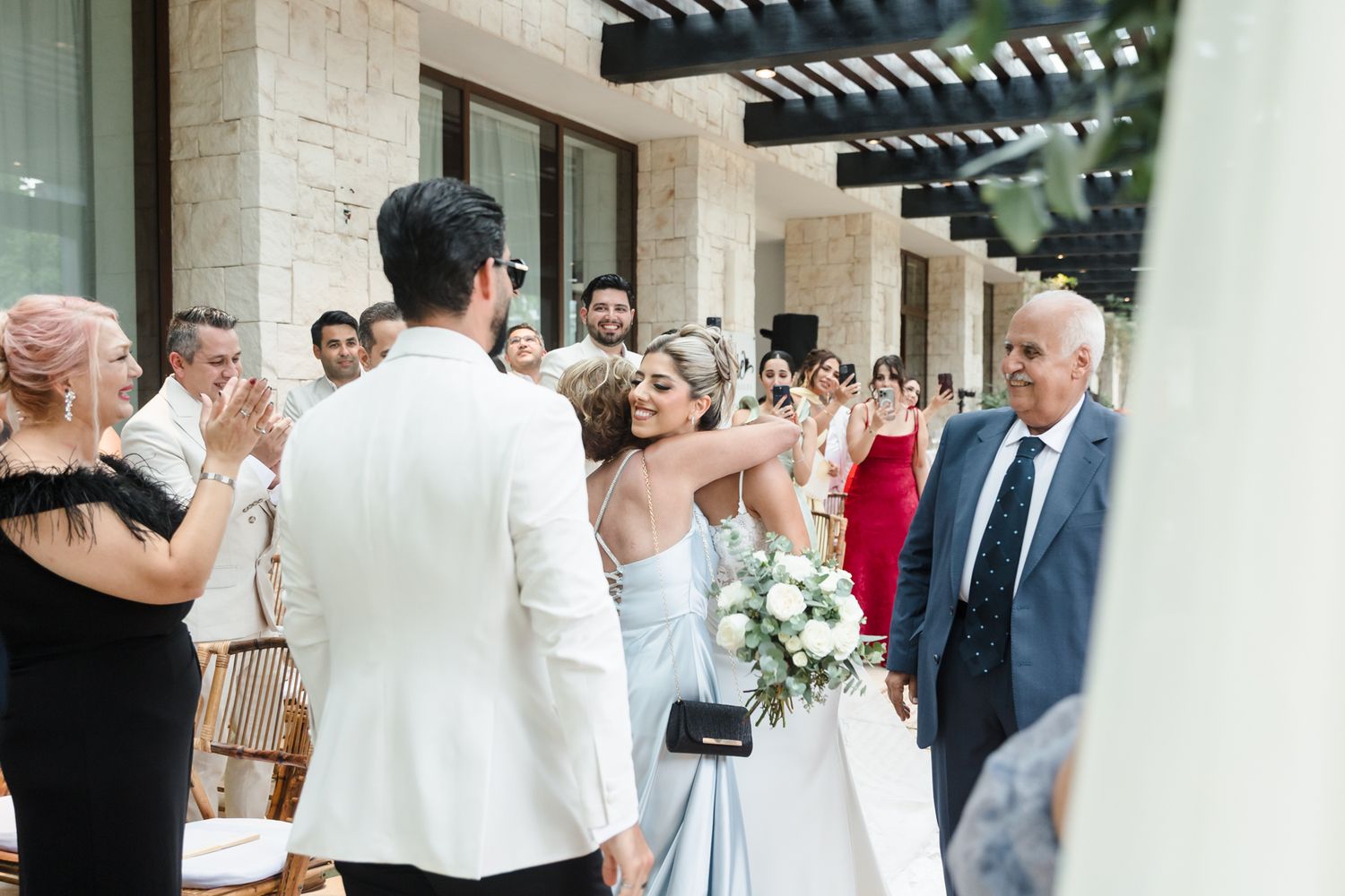 Bride in blue gown holding white bouquet greets groom at outdoor wedding ceremony with guests applauding.