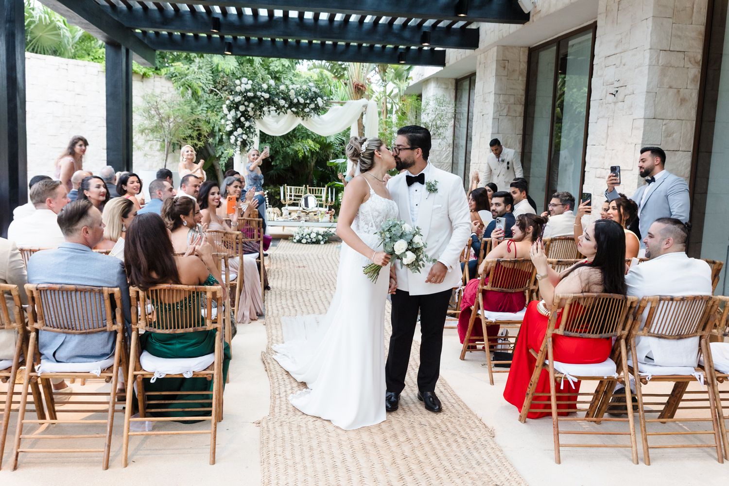 Bride and groom share a kiss at outdoor wedding ceremony with seated guests and lush greenery backdrop.