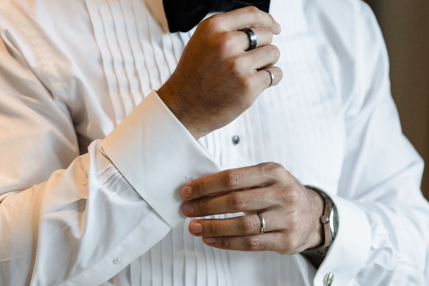 Man in white tuxedo shirt fastening silver cufflinks, wearing a black bow tie and ring, preparing for formal event.