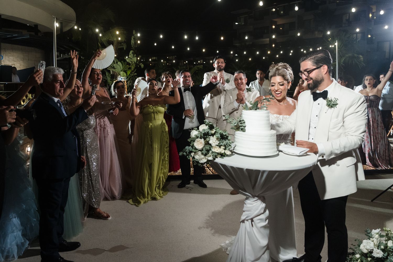 Bride and groom cut wedding cake surrounded by cheering guests at elegant outdoor evening reception.