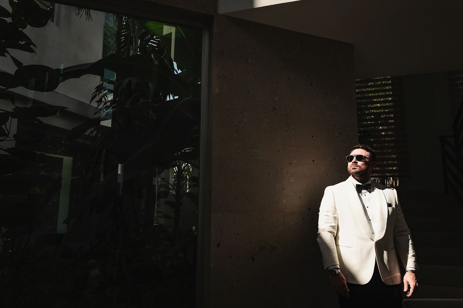 Man in white suit and sunglasses leaning against a wall in dramatic sunlight with tropical plants in background.
