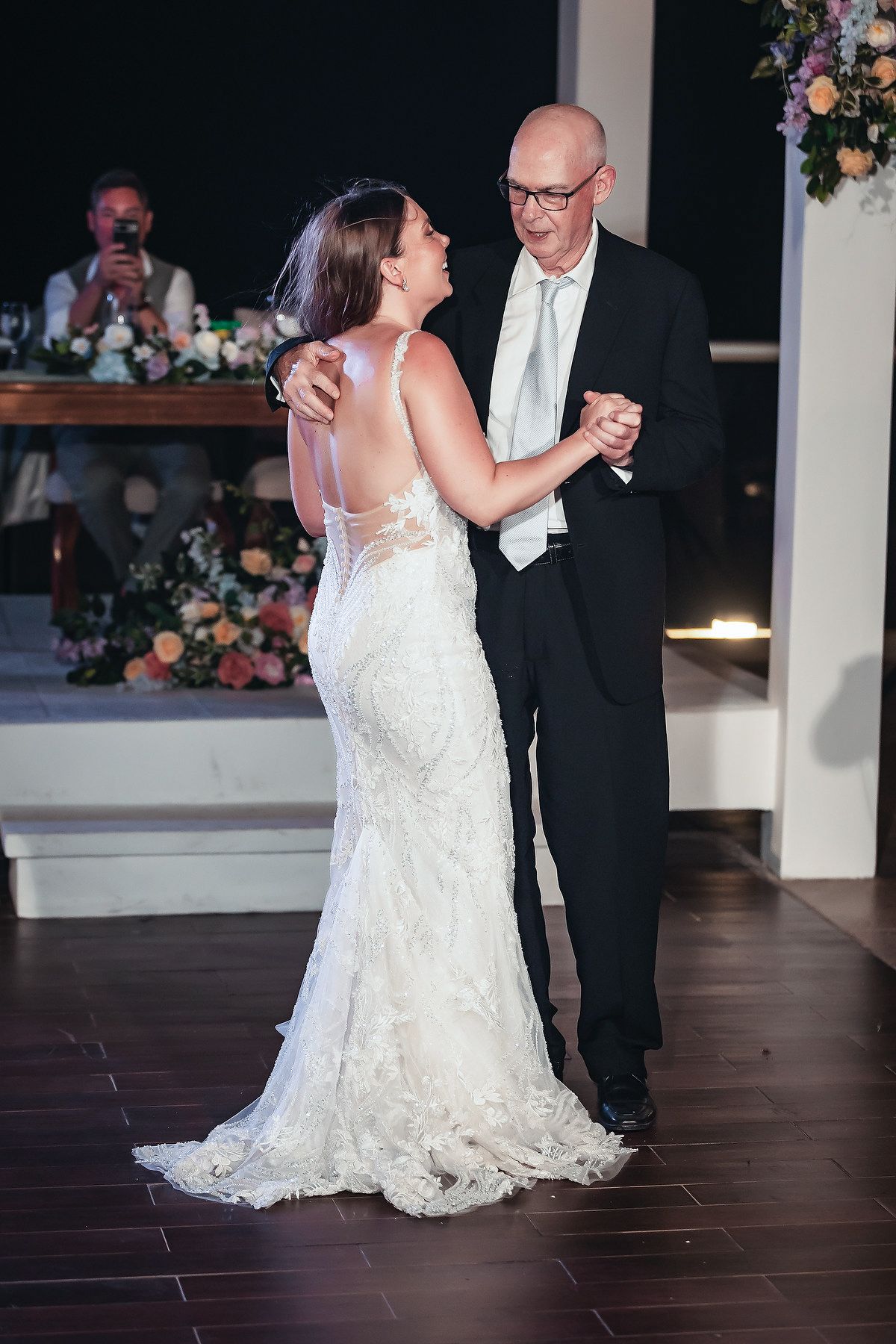 Bride in lace gown shares father-daughter dance at elegant wedding reception with floral arrangements.