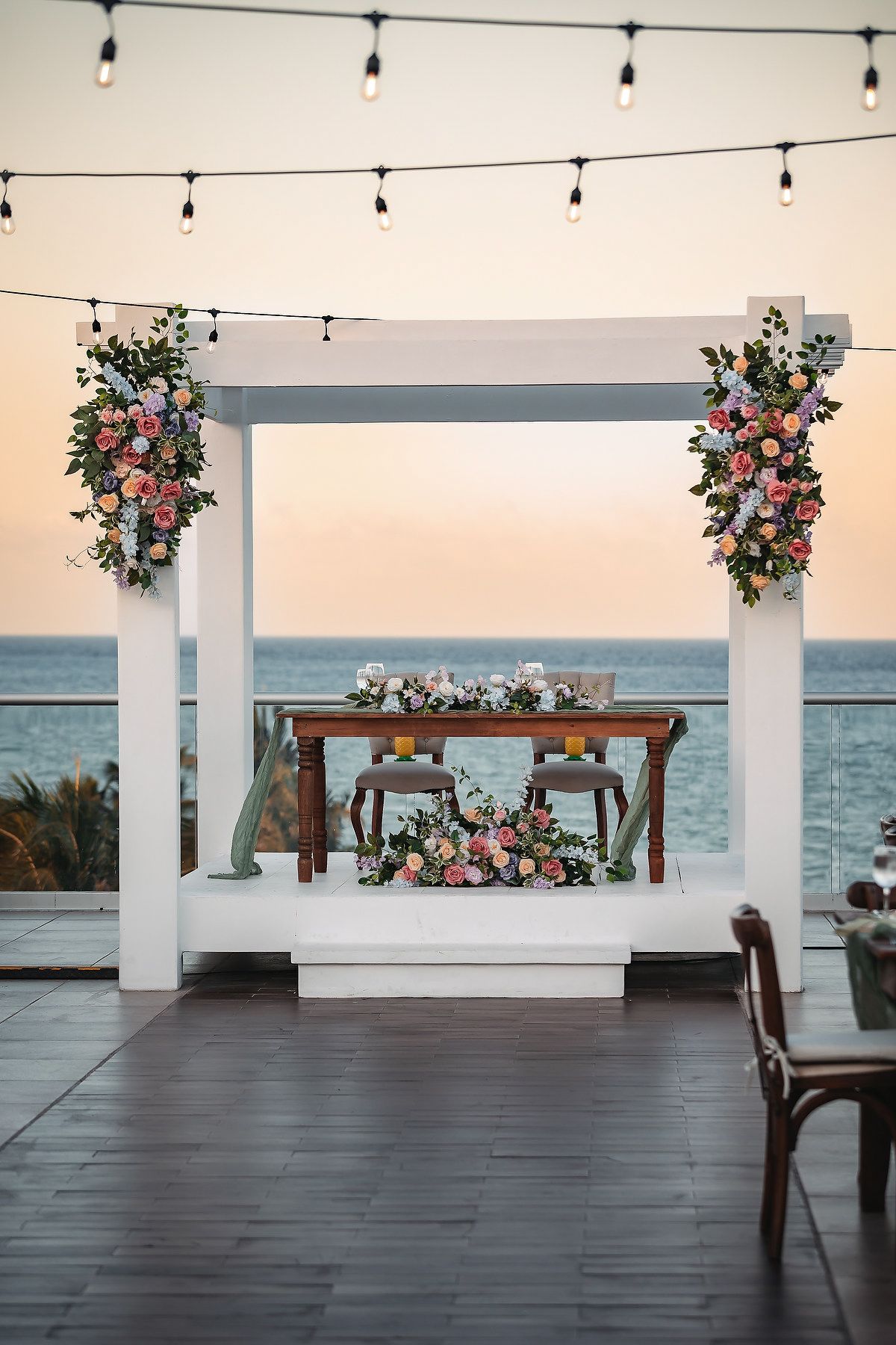 Romantic beachfront wedding sweetheart table under white floral arch with string lights at sunset.