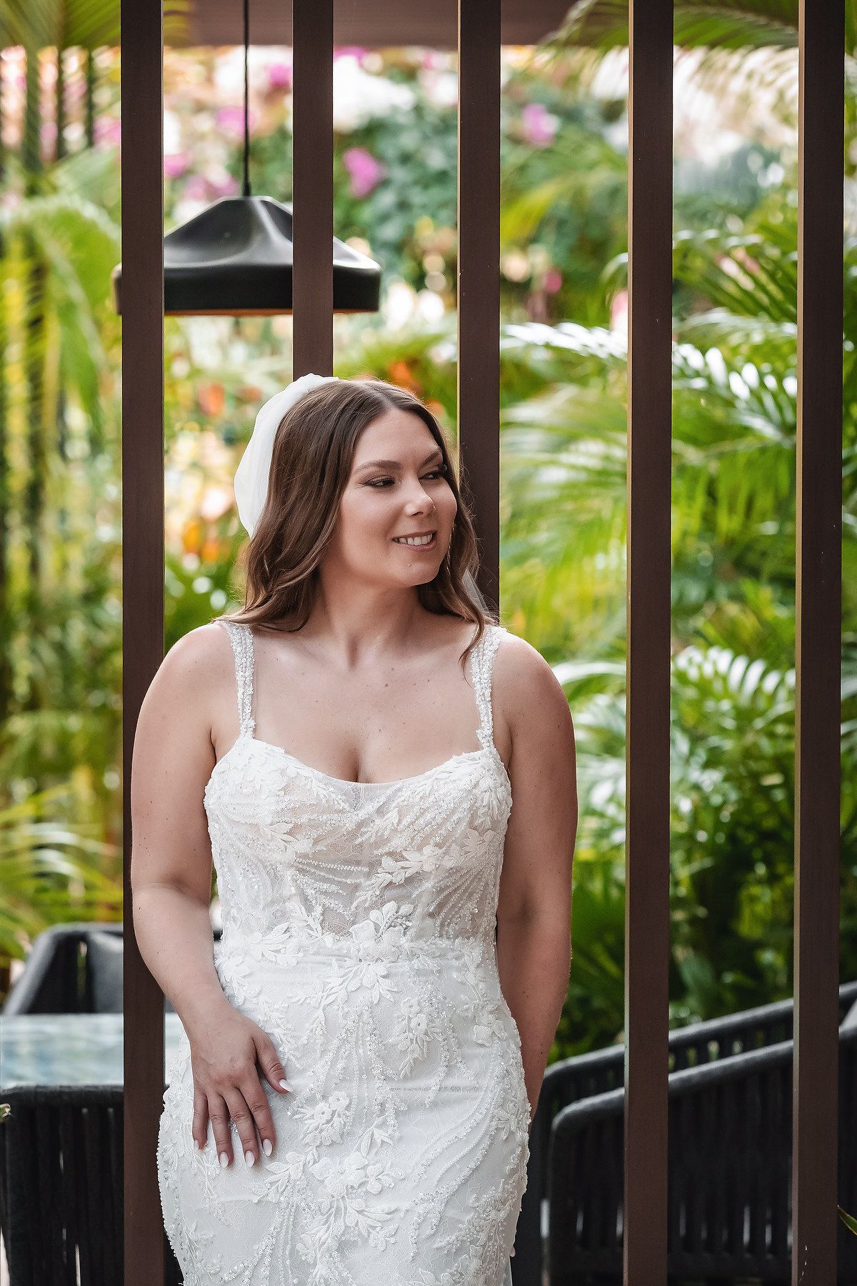 Bride in white lace gown with floral details smiles outdoors, surrounded by lush tropical greenery.