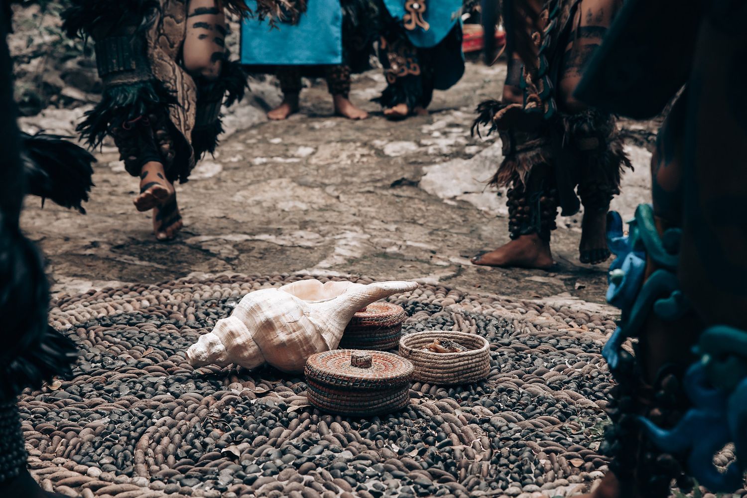 Conch shell and woven baskets placed on a decorative mat surrounded by feet of Mayan ceremony participants.