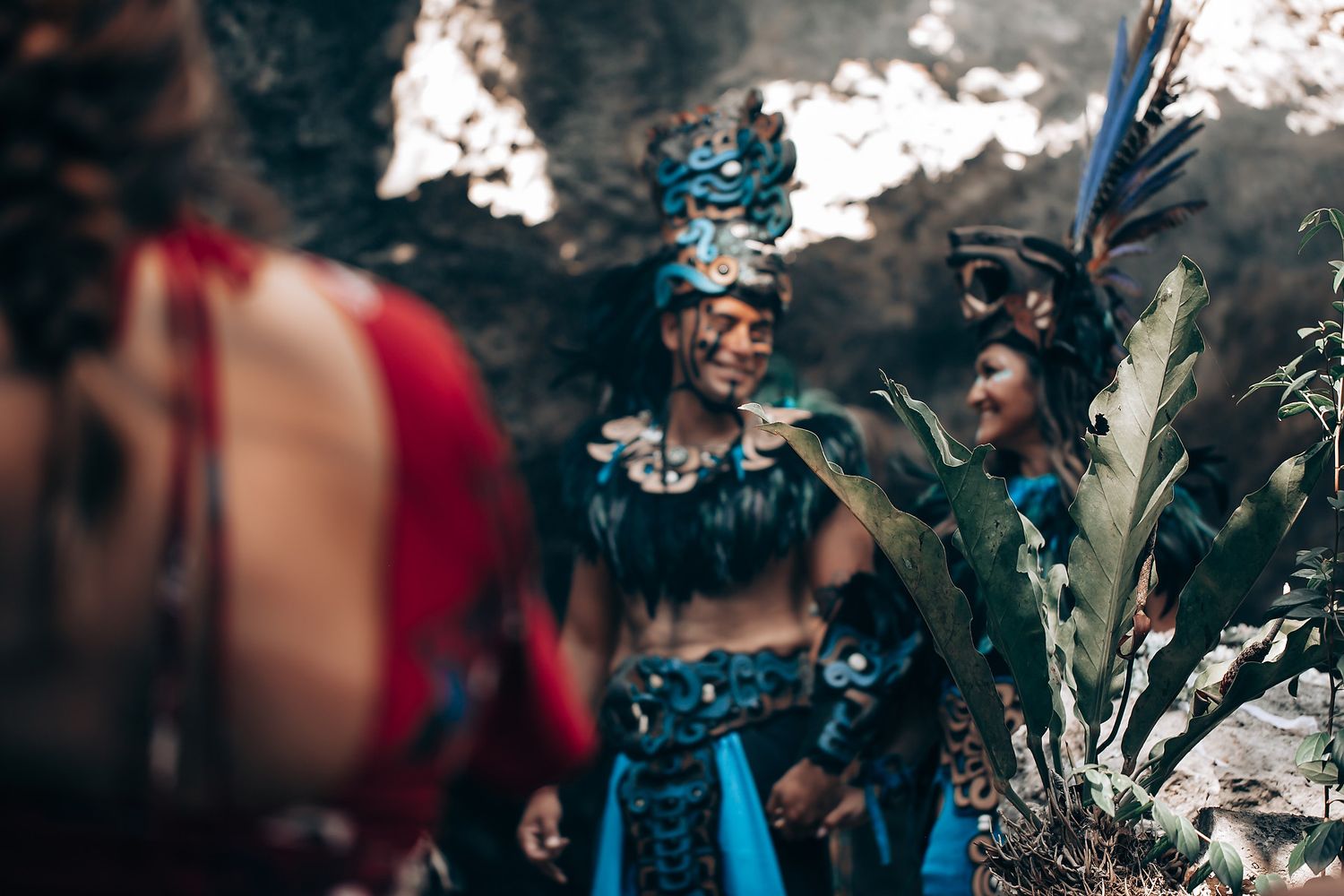 Group of performers in vibrant blue and red Mayan ceremonial costumes gathered near cave entrance.