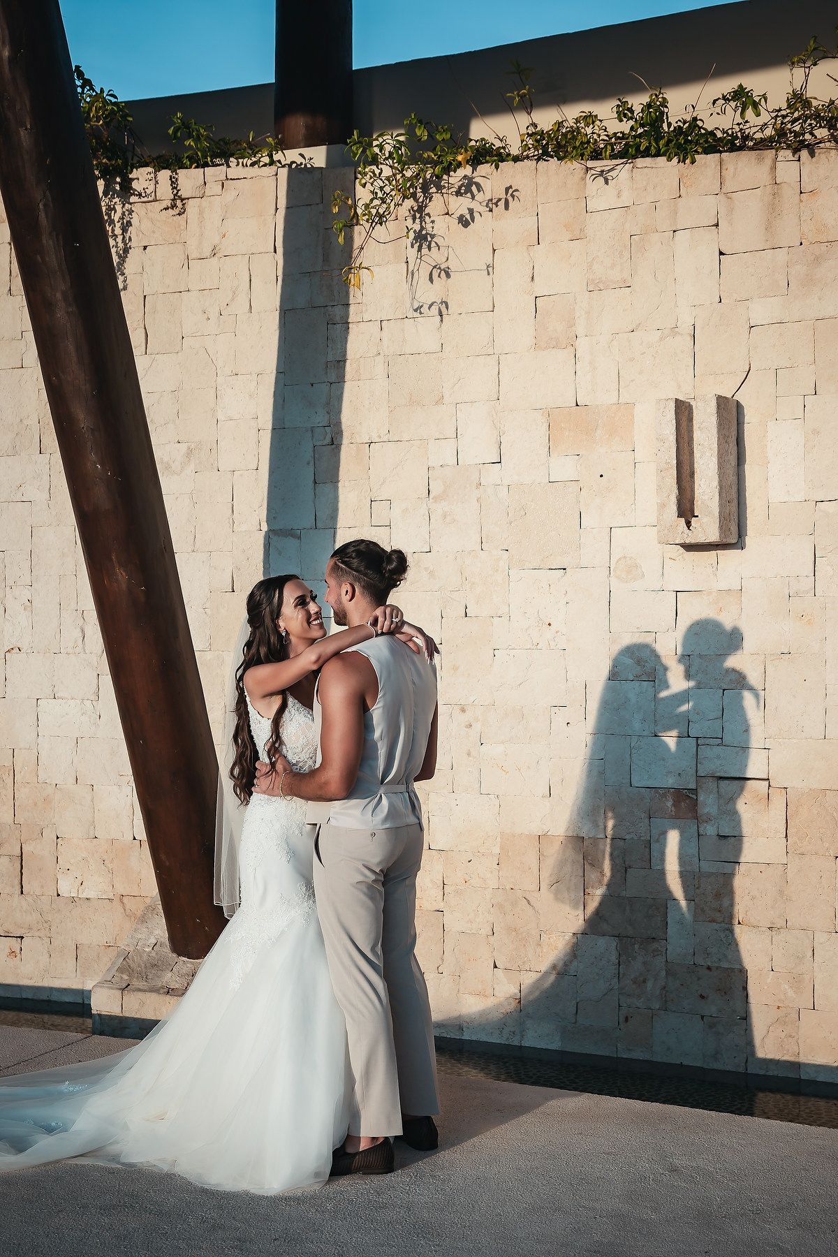 Bride and groom share a romantic embrace casting shadows on a stone wall at an outdoor wedding venue.