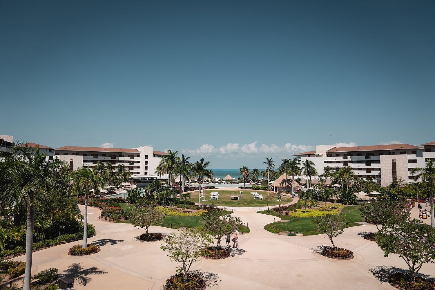 Aerial view of a luxury tropical resort with lush palm trees, manicured gardens, and ocean views under a clear blue sky.