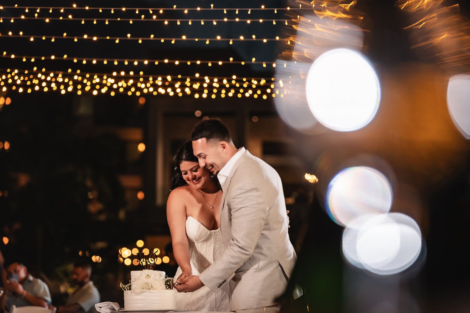 Bride and groom share a romantic moment cutting their wedding cake under warm string lights at an outdoor evening reception.