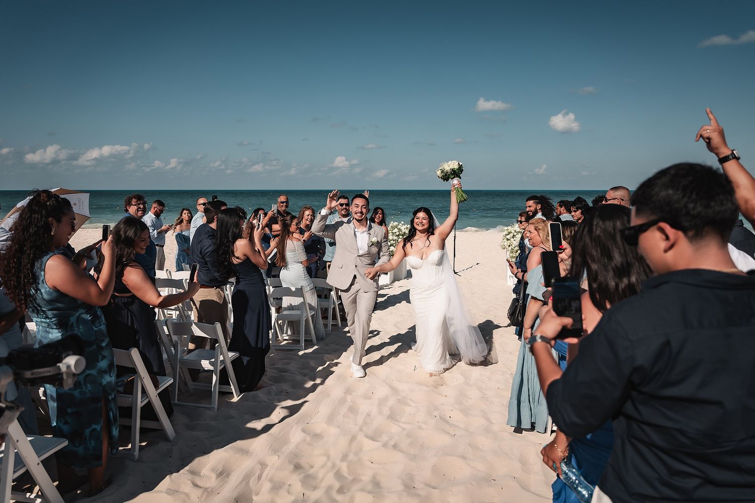 Bride and groom walk down sandy beach aisle surrounded by cheering guests under blue sky at outdoor wedding ceremony.