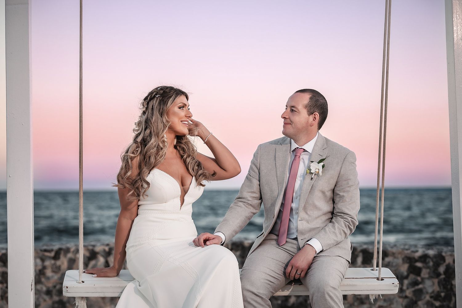 Bride and groom sitting on a white swing by the ocean at sunset, sharing a romantic glance.