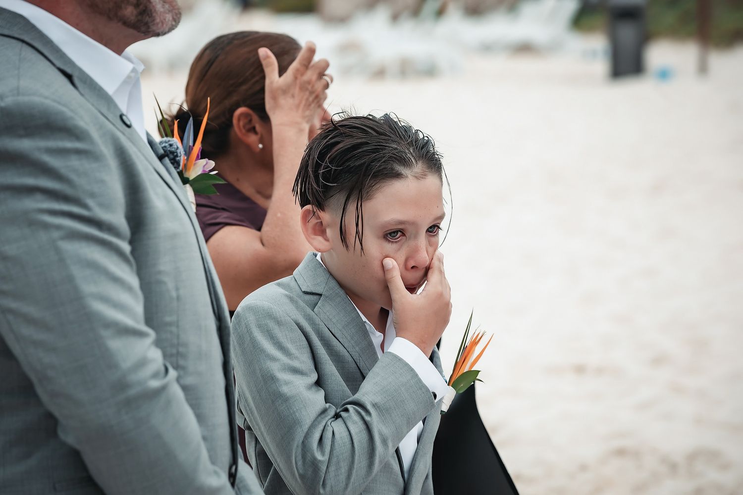 A young boy in a gray suit with a boutonniere covers his mouth emotionally during a beach wedding ceremony.