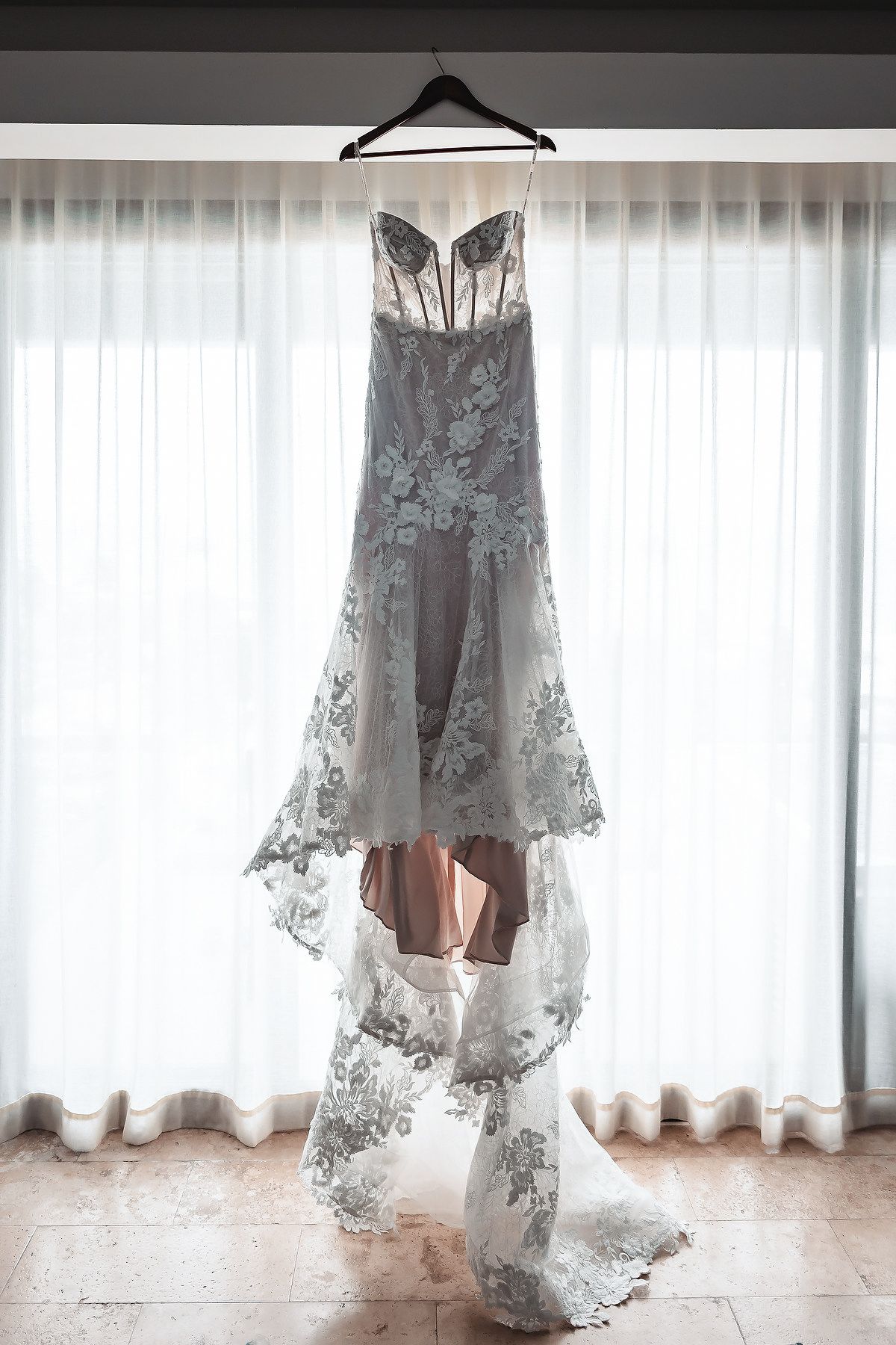 Elegant floral lace wedding gown hanging by a window.