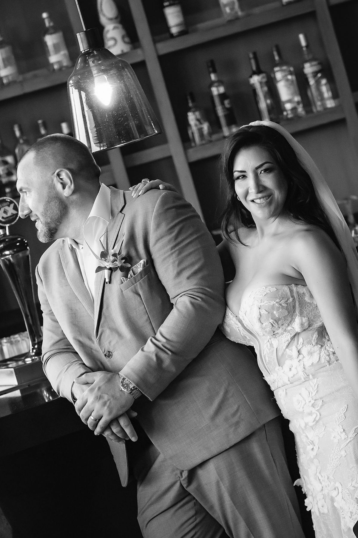 Black and white wedding photo of smiling bride in floral gown with groom in suit at a bar.