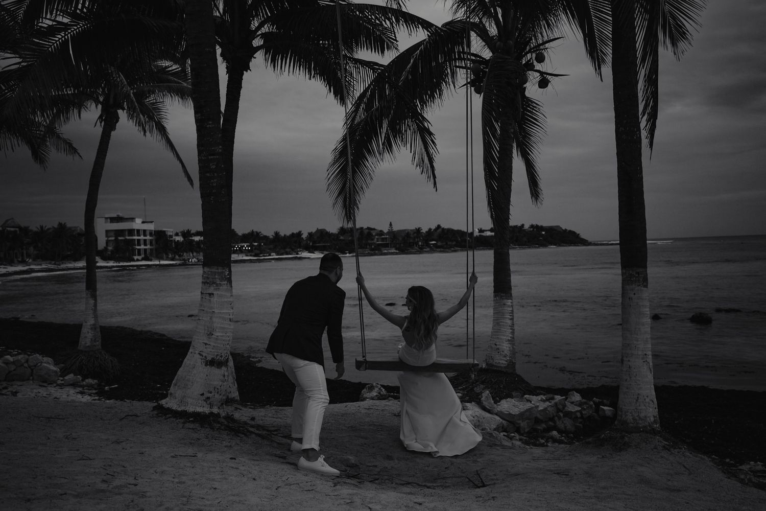 Black and white twilight photo of bride on swing with groom by palm trees overlooking ocean shore.