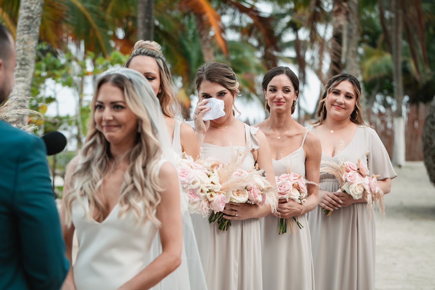 Bridesmaids in champagne dresses holding pink bouquets stand emotional during a beach wedding ceremony with palm trees.