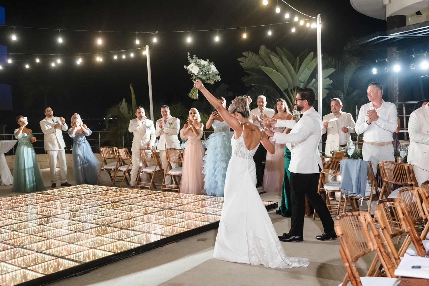 Bride and groom share first dance at outdoor wedding reception with string lights and cheering guests at night.