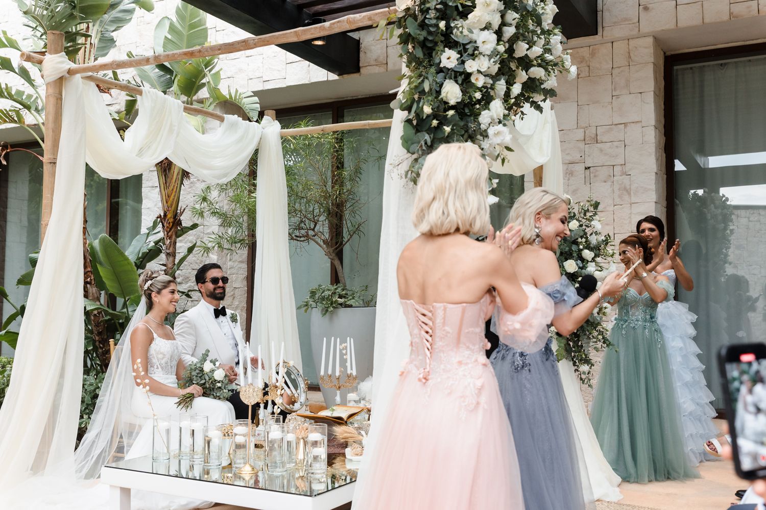 Bride embracing guest at outdoor wedding with floral arch, white draping, and bridesmaids in pastel gowns.