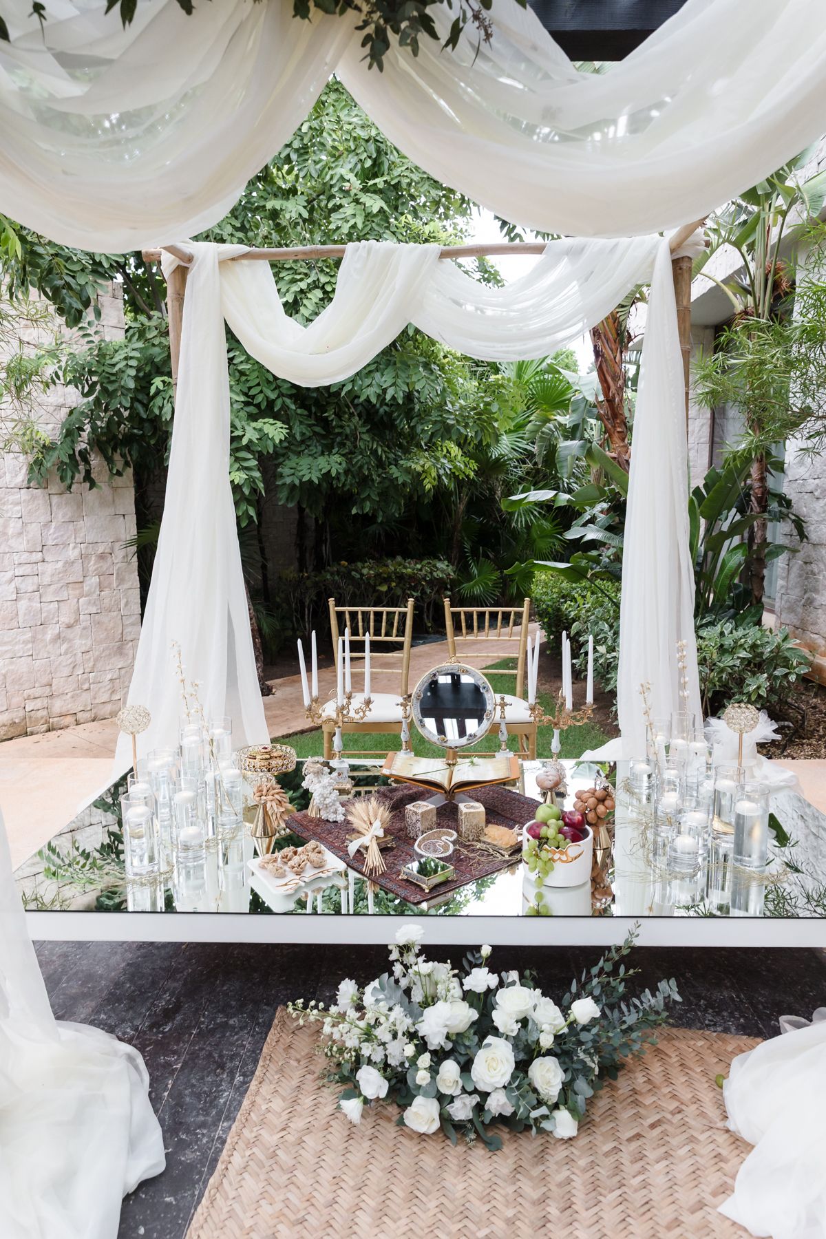Elegant outdoor wedding ceremony setup with white draped canopy, mirrored table, candles, and lush greenery backdrop.