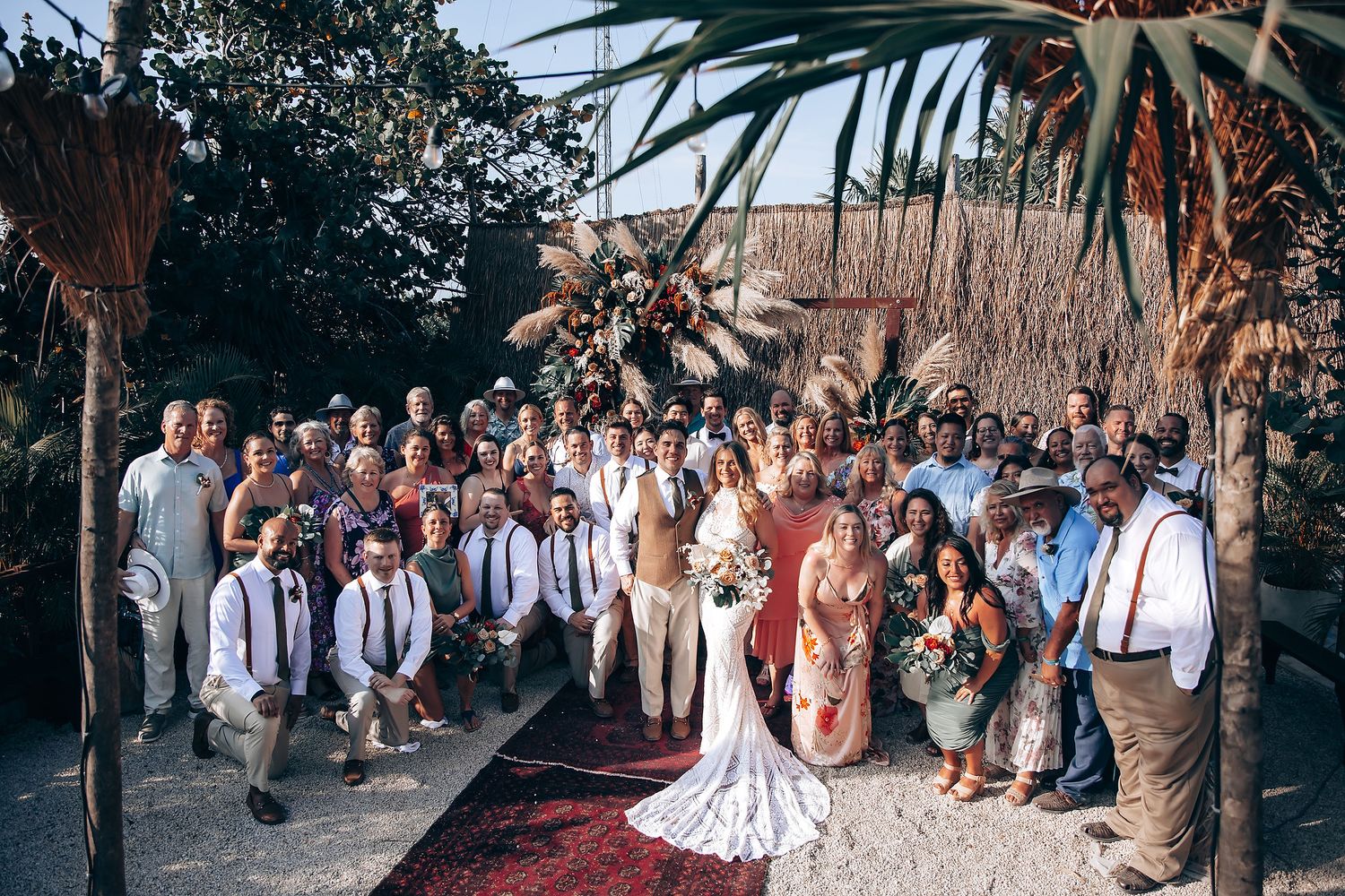 Outdoor wedding celebration with guests seated beneath tropical palm fronds and pampas grass decorations.
