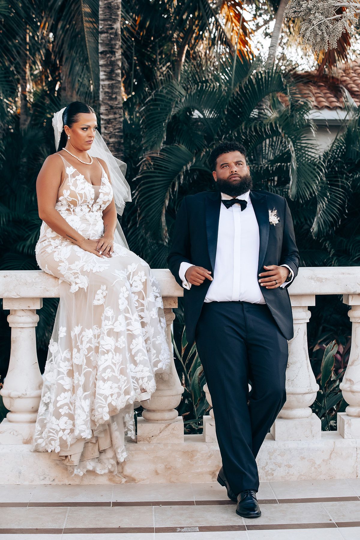 Romantic wedding couple sitting on ornate white balustrade surrounded by palm trees.