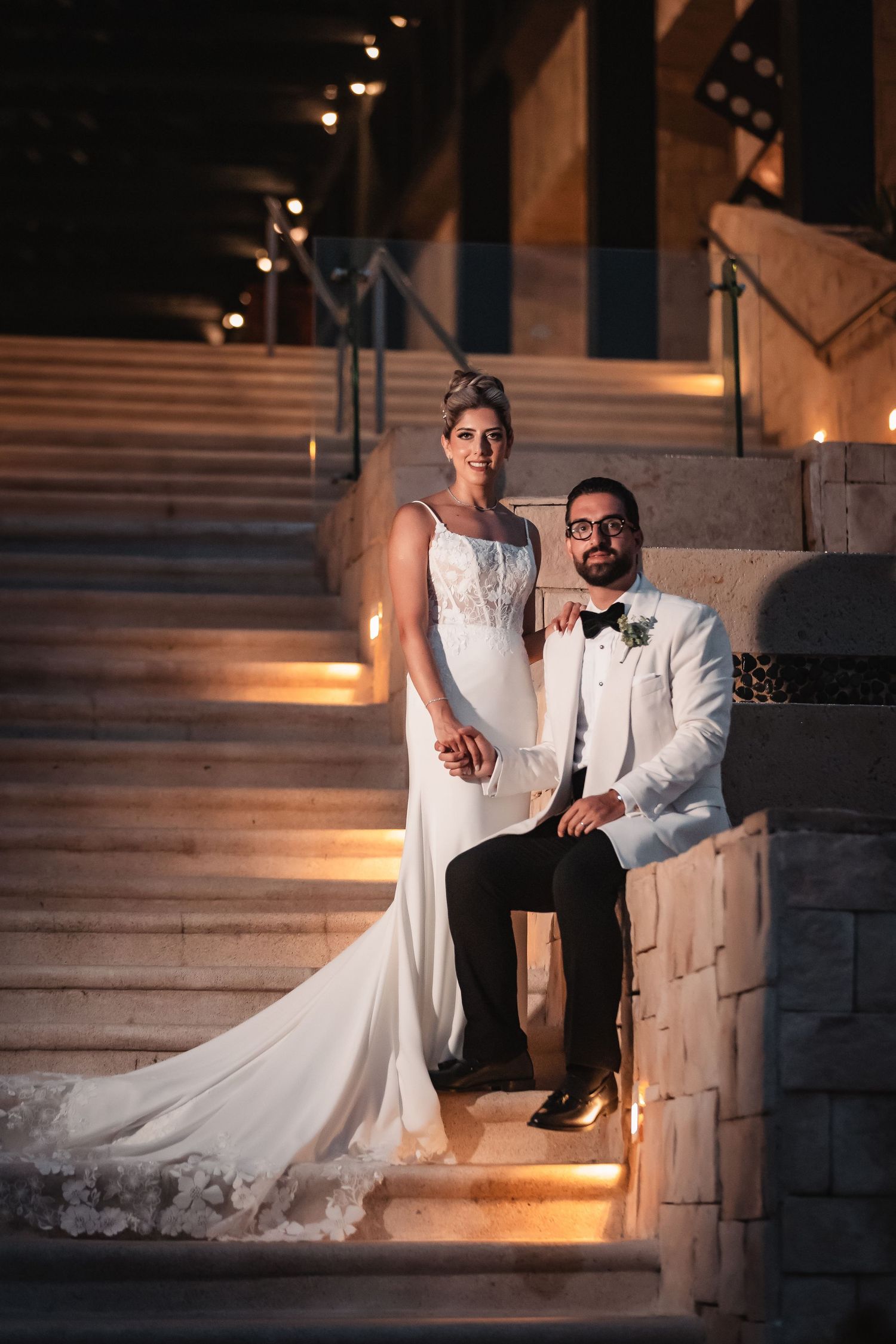 Wedding couple posing on illuminated stone steps at dusk in elegant attire.