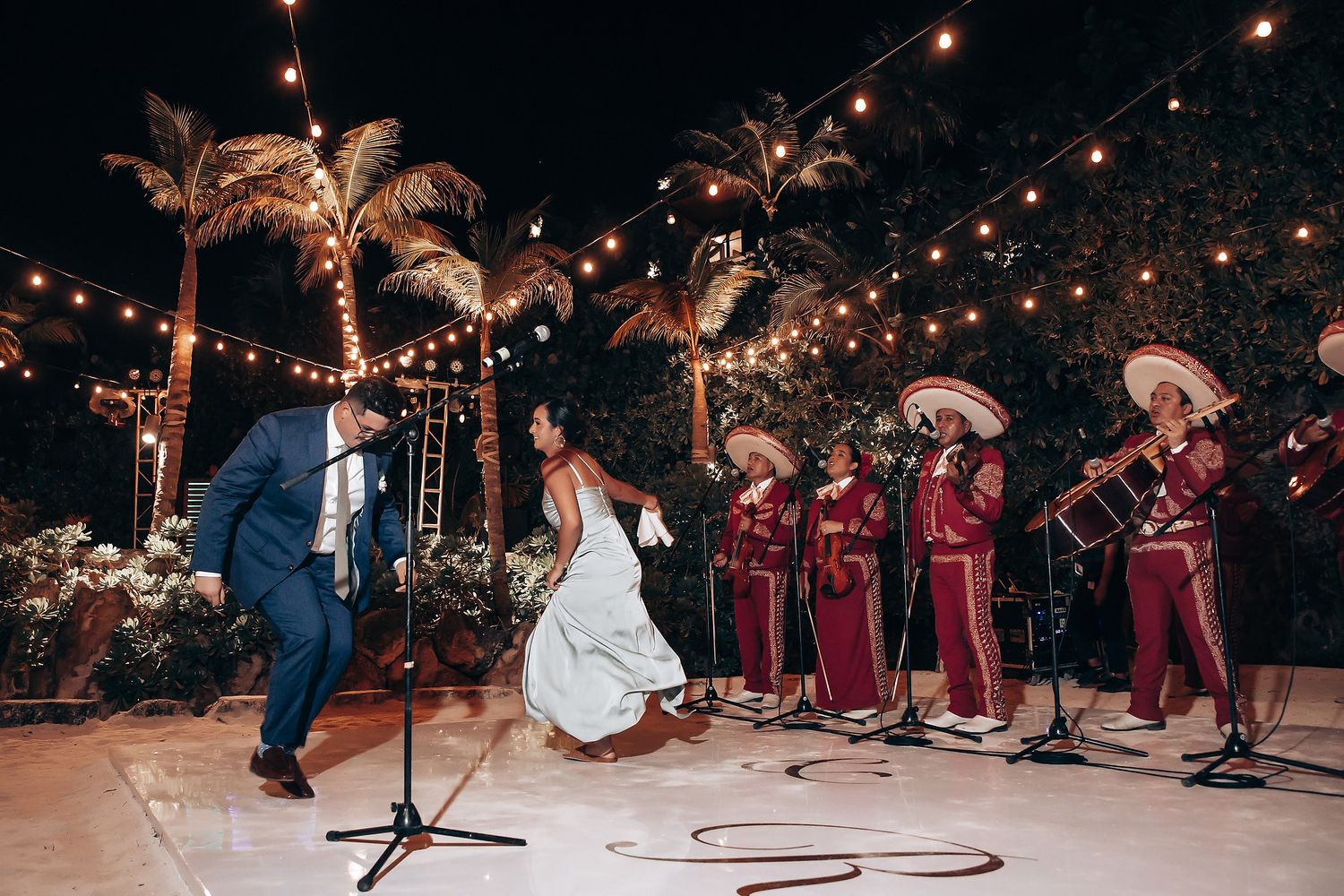 Wedding party dances under string lights at an outdoor tropical reception venue.