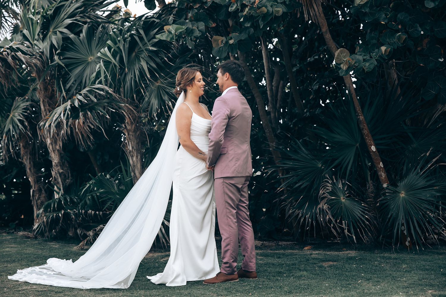Romantic wedding couple embracing in a lush garden setting with palm trees in the background.