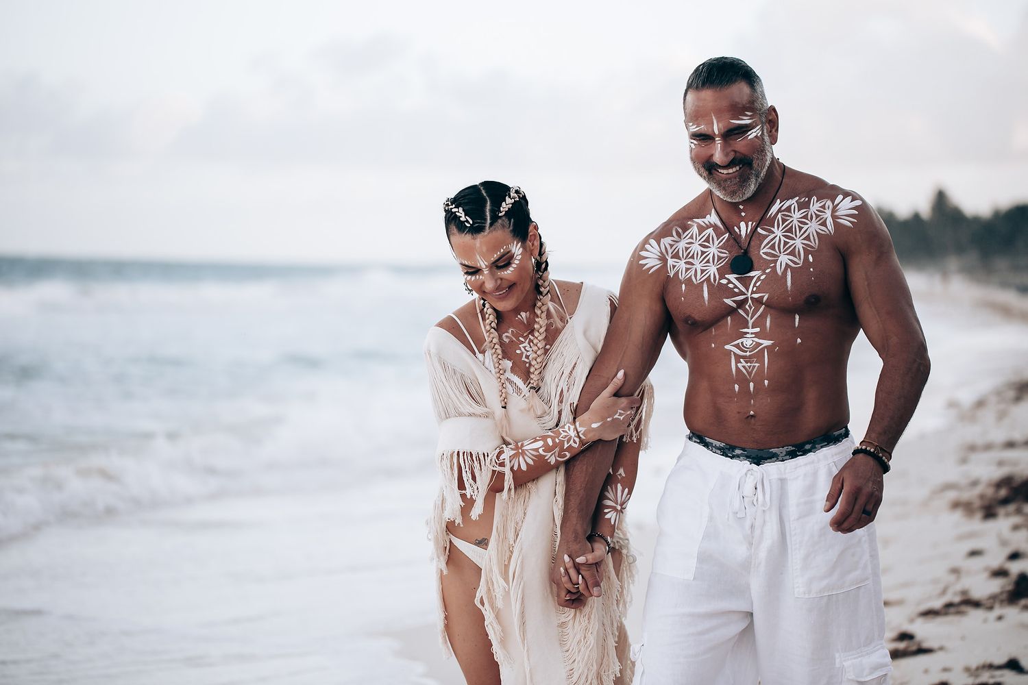 A couple in bohemian beach wear with white body paint poses on the sand.