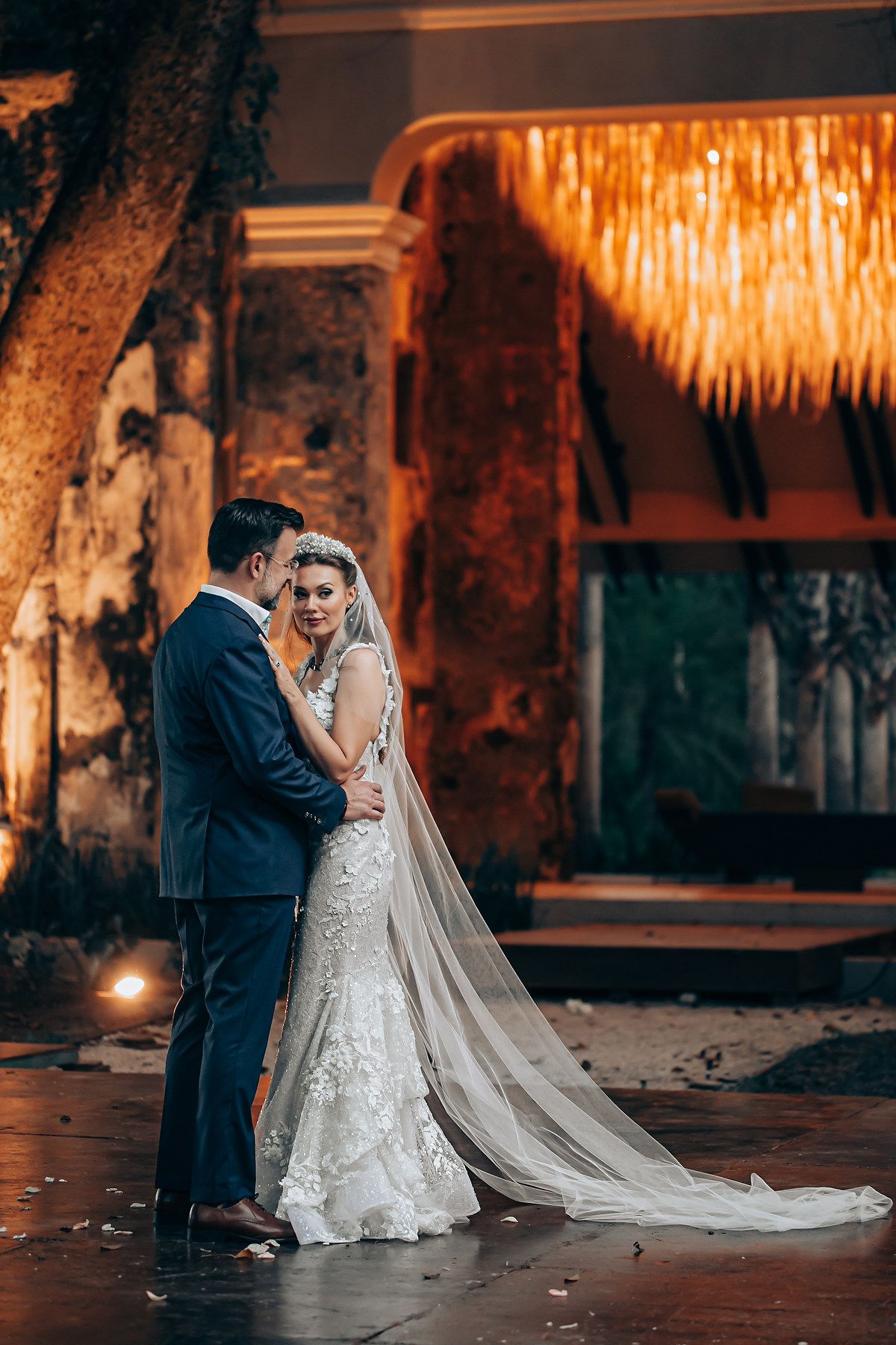 Romantic wedding couple embracing under warm lighting with a dramatic cathedral veil trailing behind.