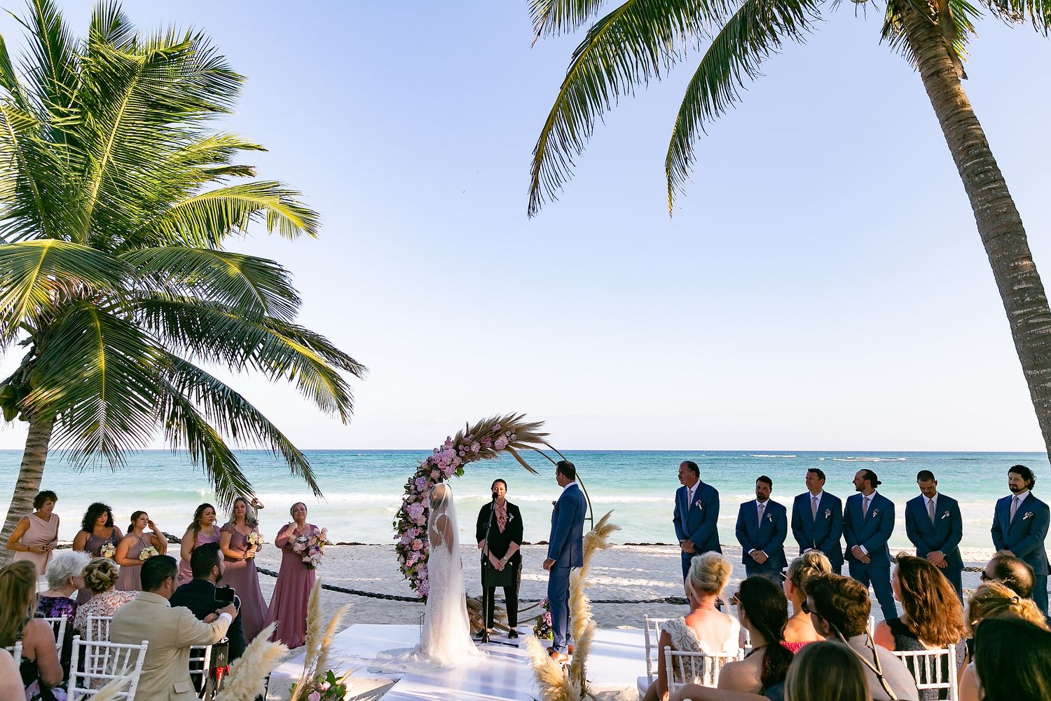 A tropical beach wedding ceremony setup with palm trees swaying in the breeze and chairs arranged on white sand.