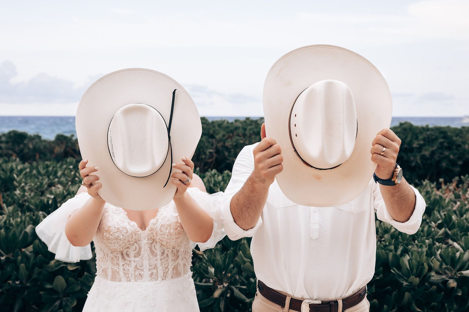 Two people hold wide-brimmed white hats against a scenic mountain backdrop in romantic outdoor setting.