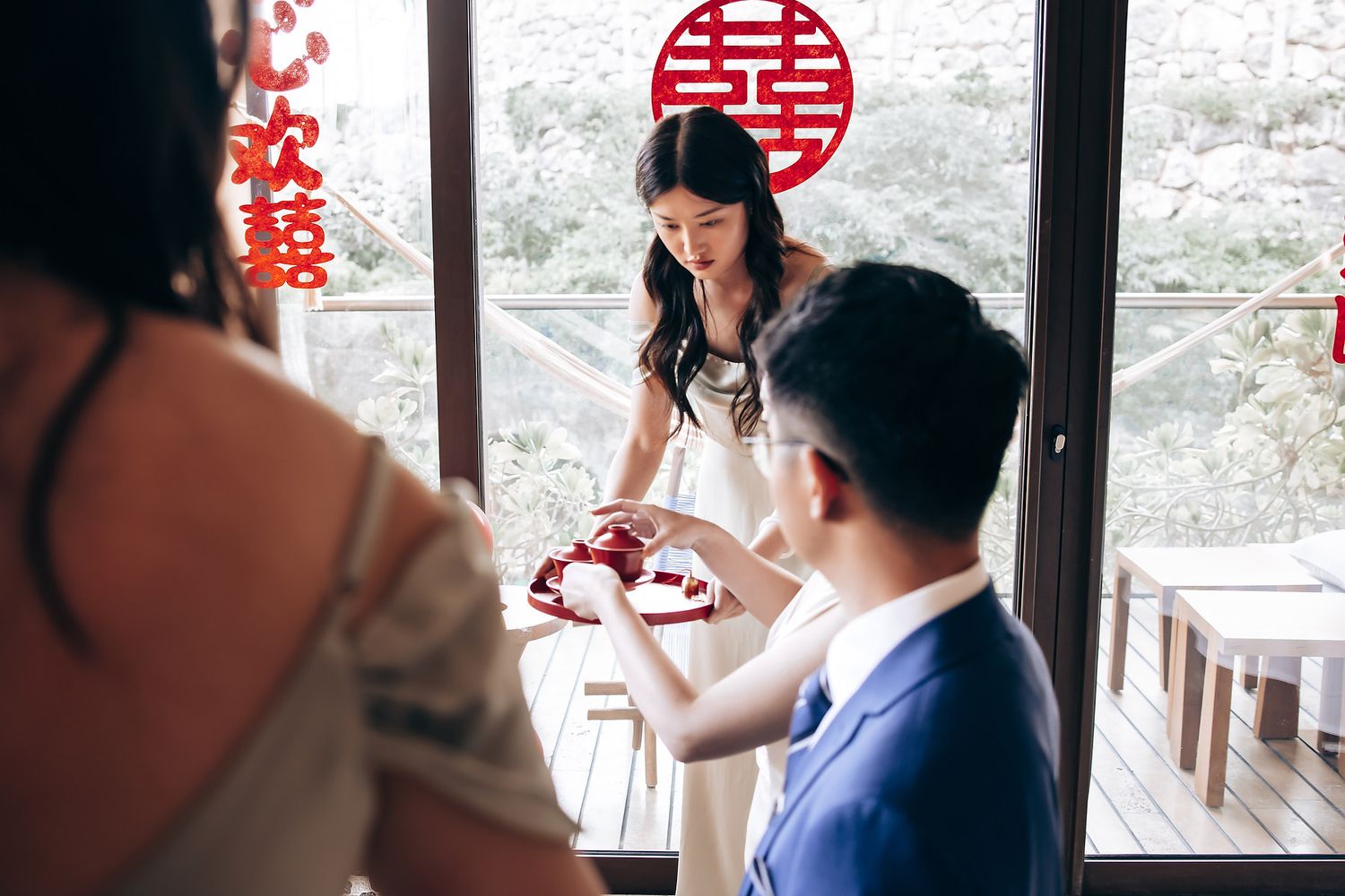 Tea ceremony participants seated by a window with traditional Chinese decorations and symbols.