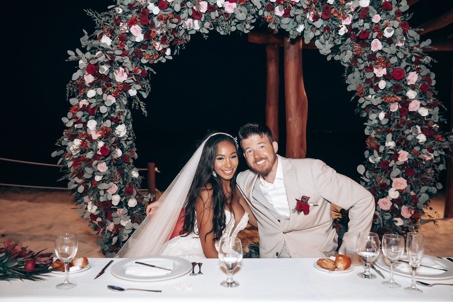Wedding reception table decorated with red and white floral arrangements under elegant arch.