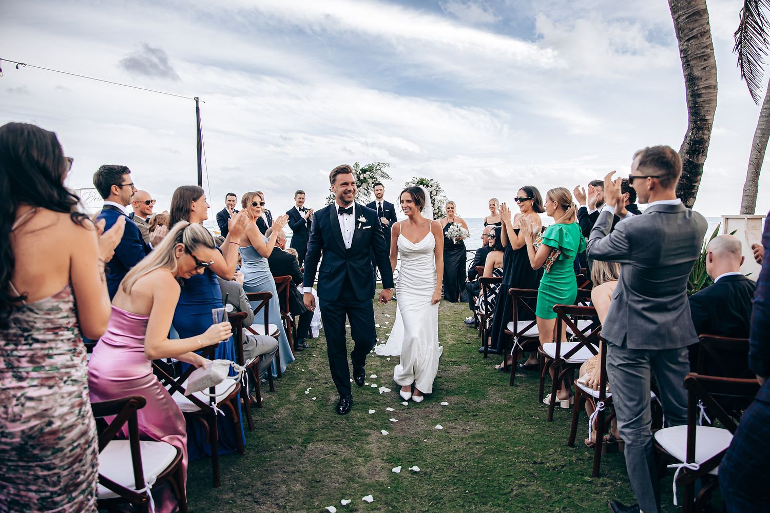 Outdoor wedding ceremony setup with rows of white chairs and tropical landscaping by the ocean.