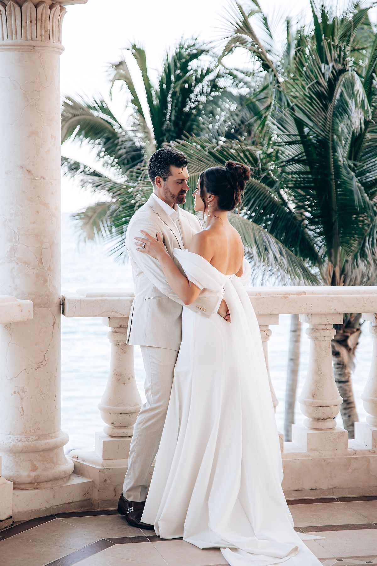 A newlywed couple embraces on a tropical balcony with palm trees and white columns in the background.