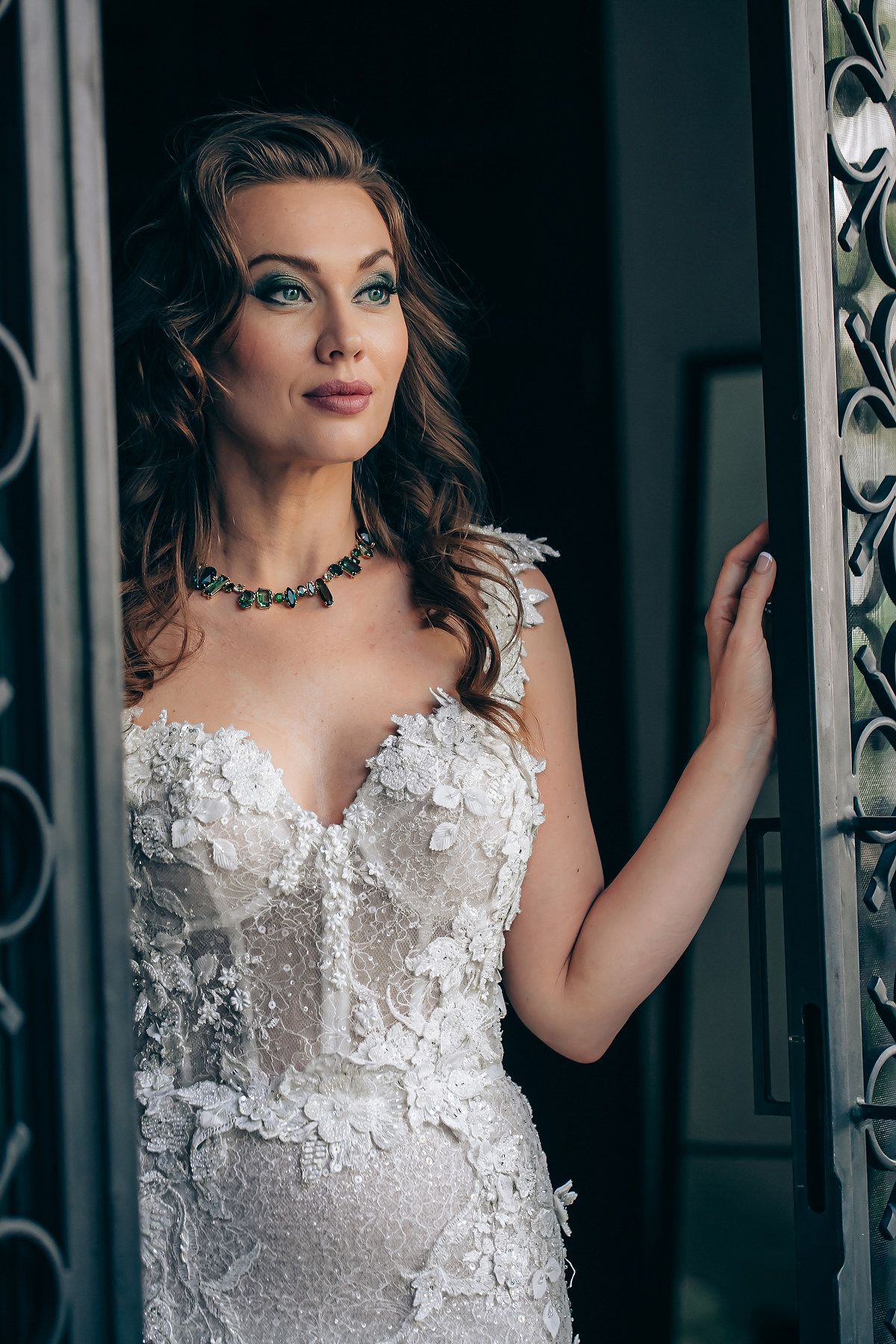 Bride in an ornate white lace wedding gown with sweetheart neckline and emerald necklace poses elegantly by doorway.