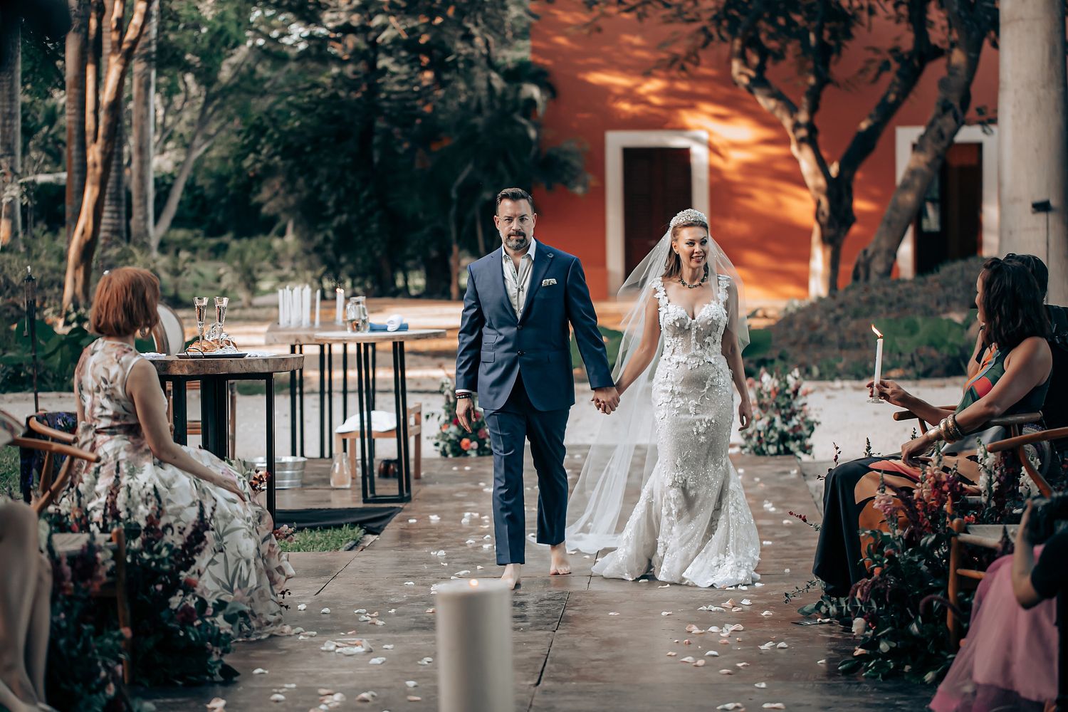 Wedding couple walks together through a garden pathway during sunset while guests shower them with flower petals.