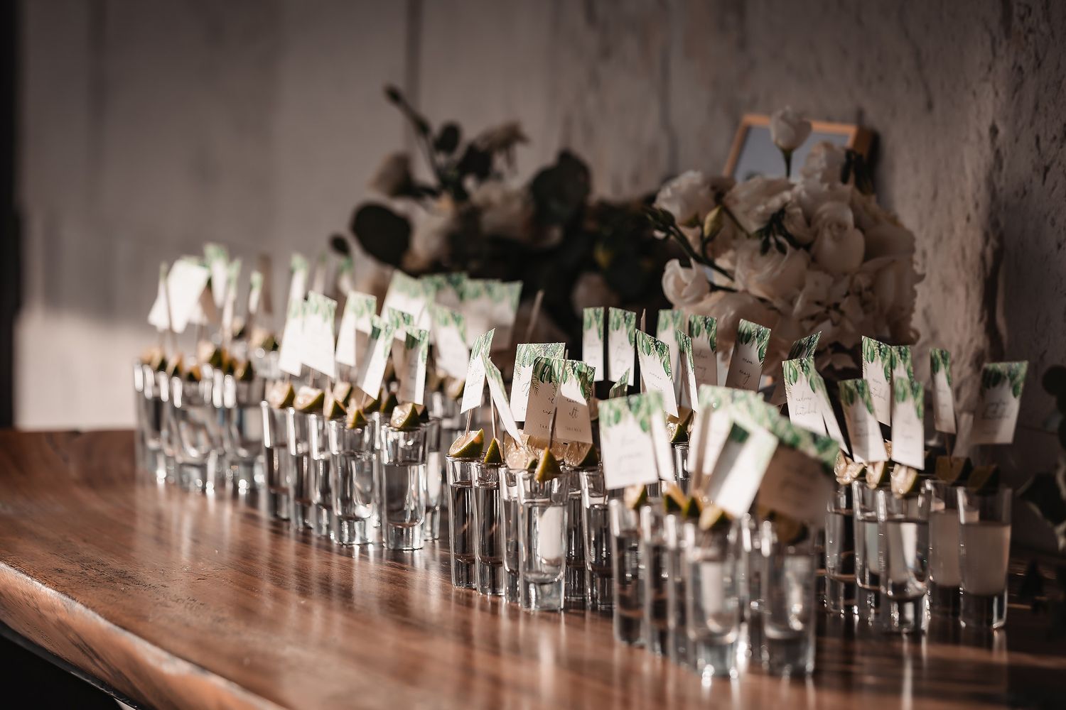Wedding seating cards arranged with greenery in crystal vases on dark wooden surface.