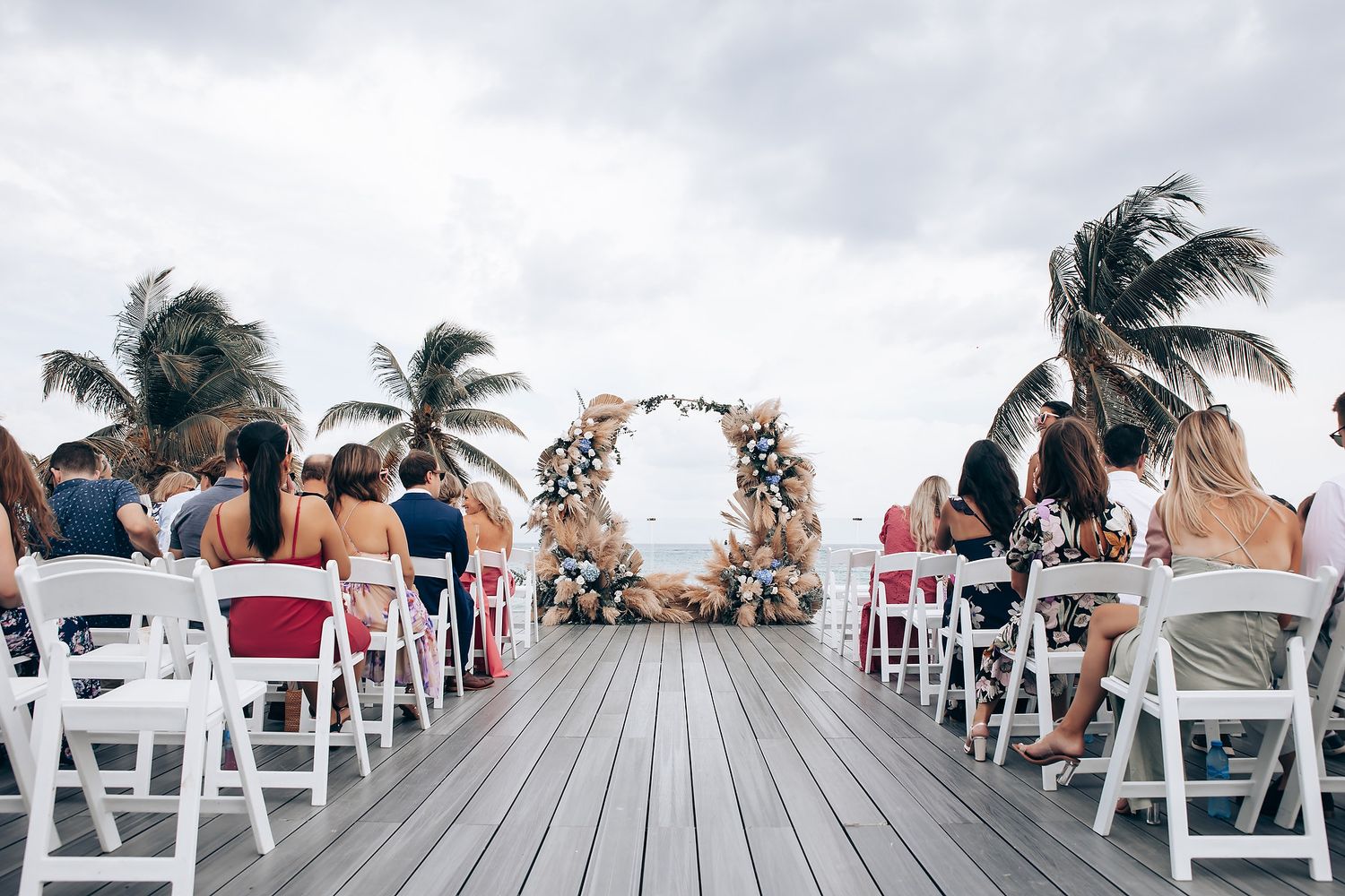 Wedding guests seated in white chairs on a wooden deck overlooking the ocean with palm trees swaying in the breeze.