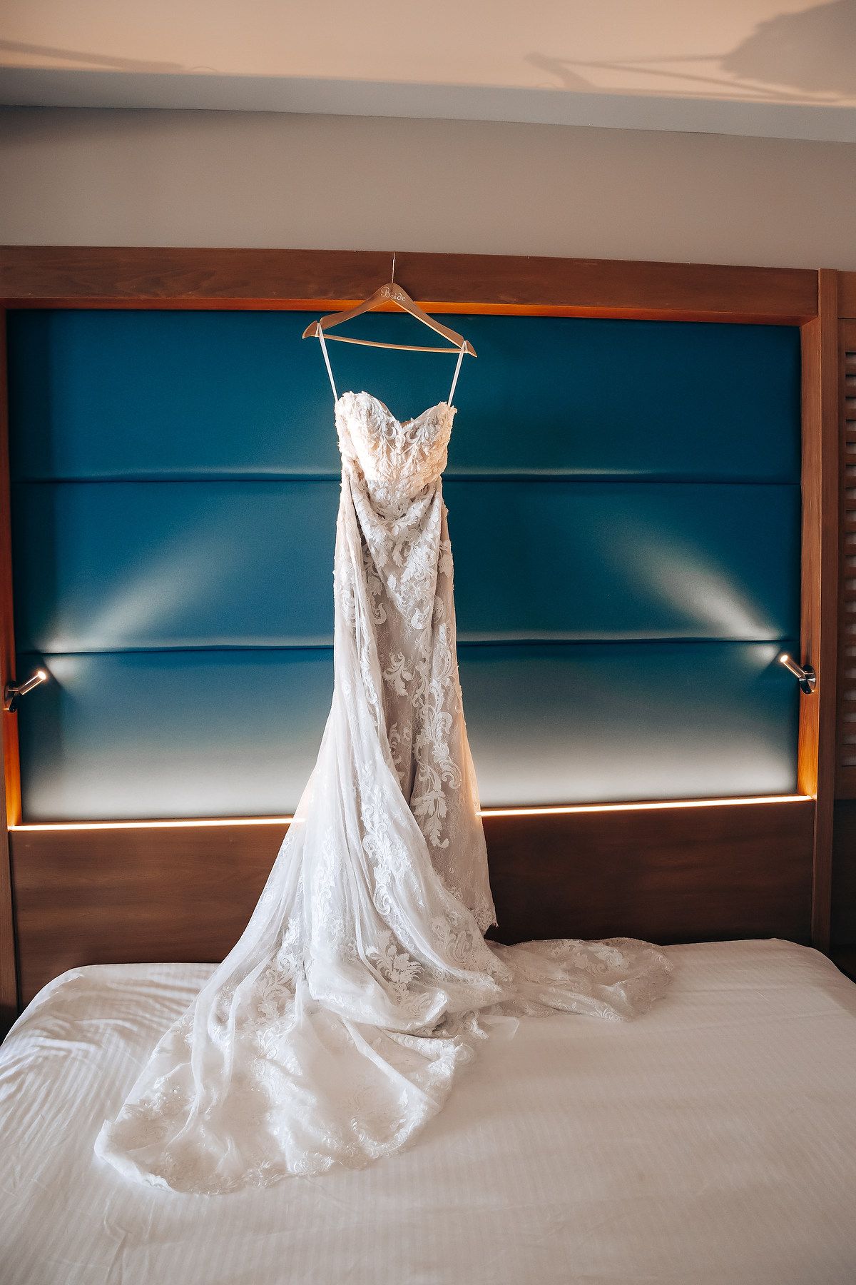 An elegant white lace wedding dress hangs on display against a modern blue and brown hotel room headboard.