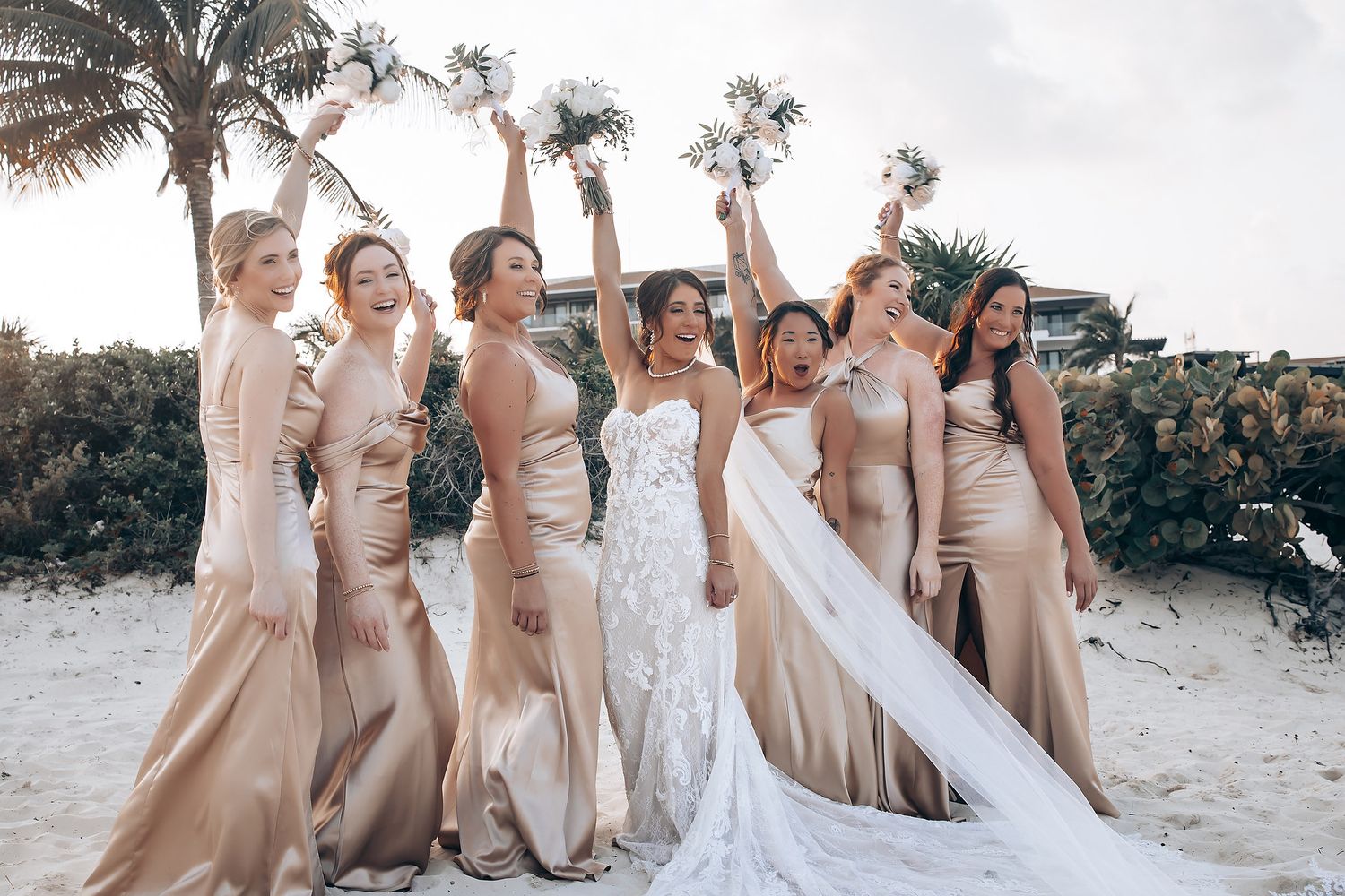 Wedding party in champagne colored dresses poses together on a sandy beach.